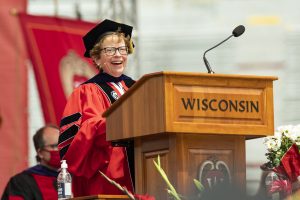 Becky Blank speaks at a podium while wearing red and black graduation regalia.