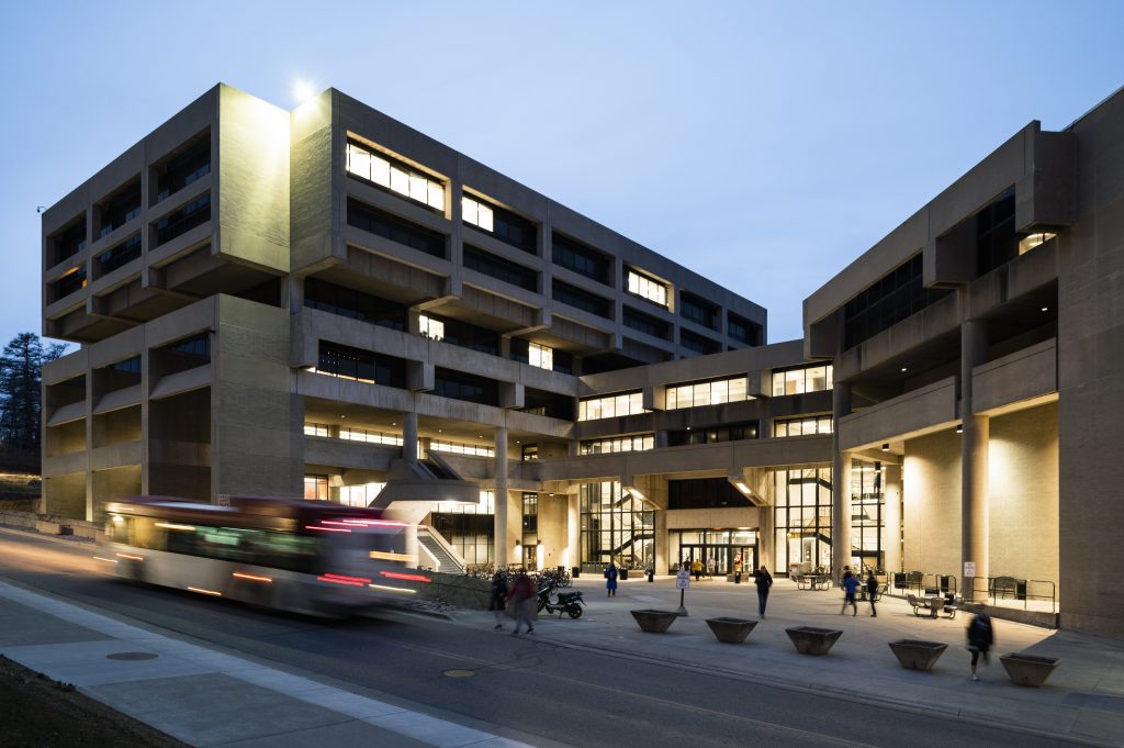Pedestrians and a campus bus passing a lit Helen C. White College Library at dusk.
