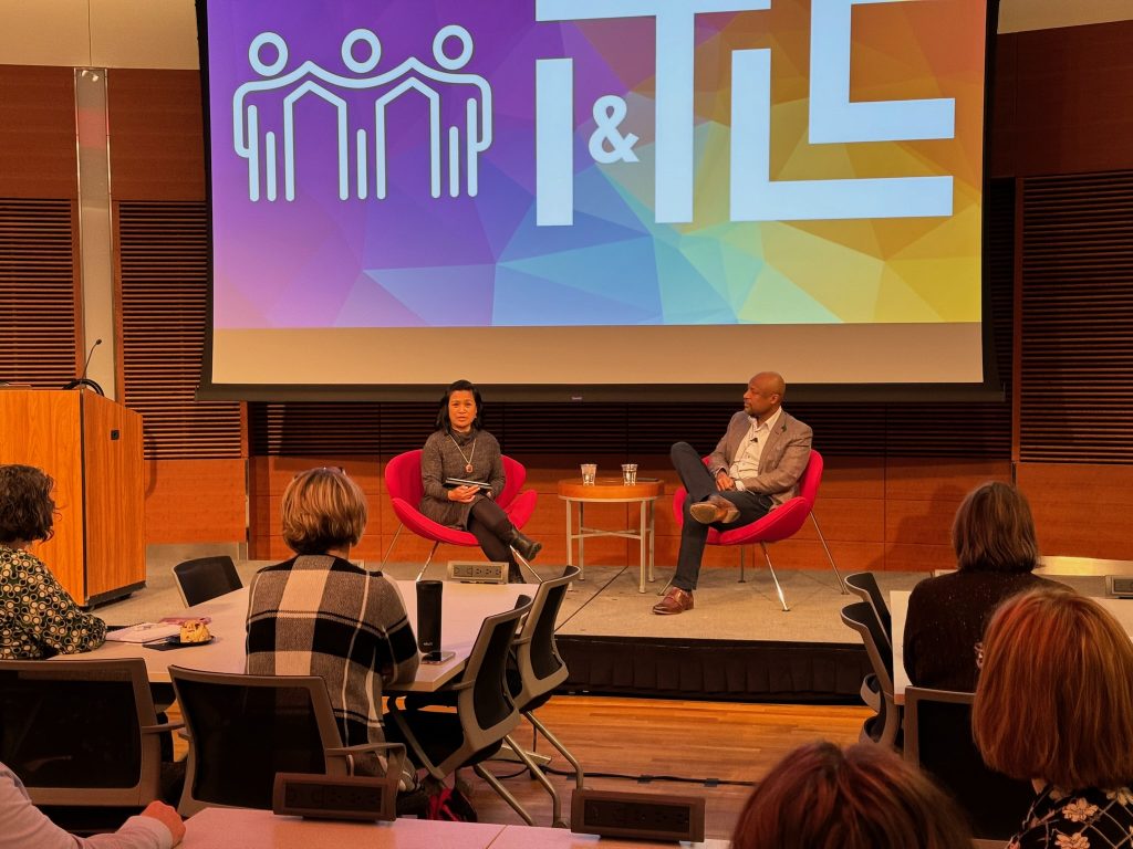Two people sit in chairs on stage in front of an audience with a slide projected behind them.