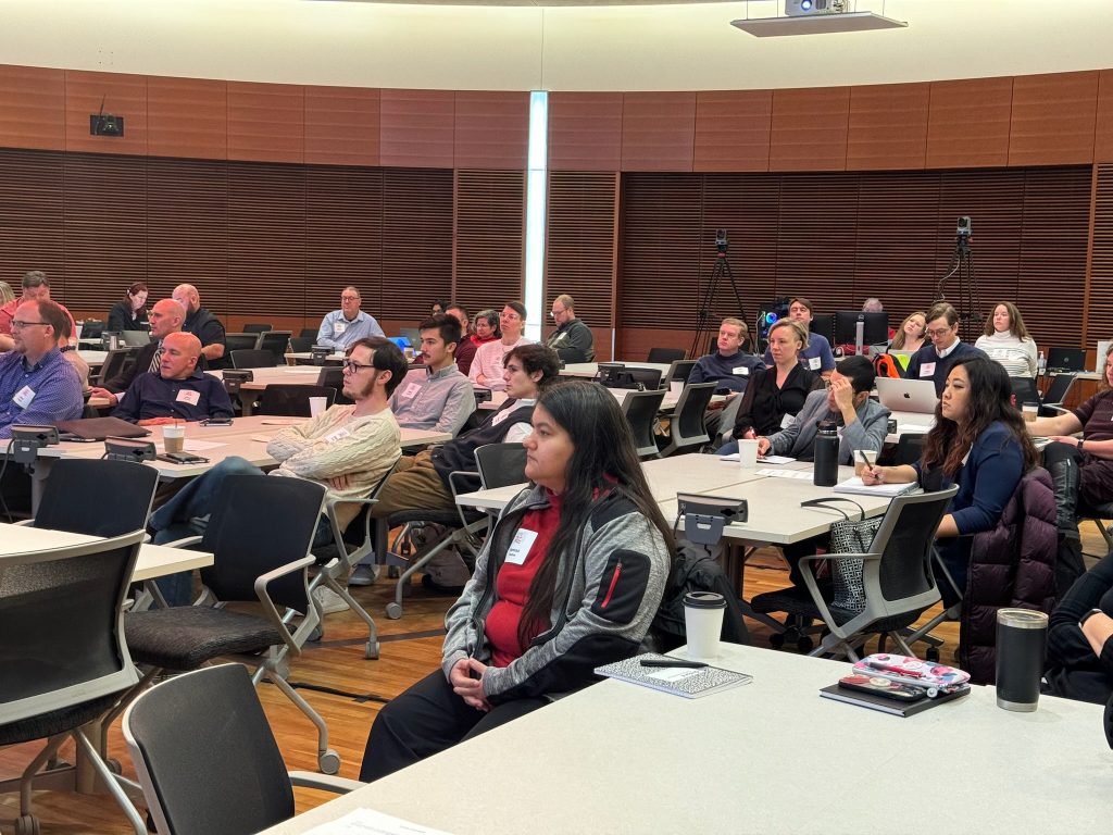 Dozens of people sit in chairs watching a speaker in a large conference room
