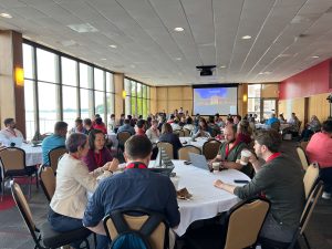 Conference attendees fill the room and sit at round tables, talking and networking before the conference begins.