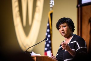 Mae Jemison speaks into a microphone on stage with a US flag and the UW crest behind her.