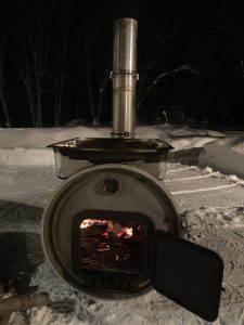 A large steel drum laying on its side with wood burning inside and evaporator trays resting on top sitting in a plowed snowy area at night.