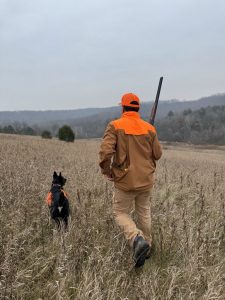 Man with a hunting rifle and a dog both wearing orange hunting gear walk through a field of high grasses.
