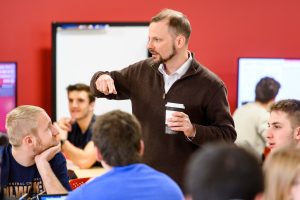 A man stands gesturing in a classroom while students watch.