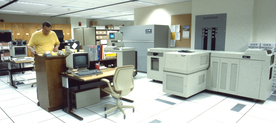 A man in a yellow shirt works in a large room full of computers and printers.