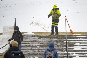 Person in hi-vis snowsuit applies salt to cleared staircase after snowfall as multiple individuals in winter clothing walk by.