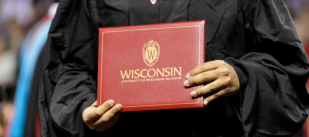 Person wearing black robes holding a red UW–Madison diploma case